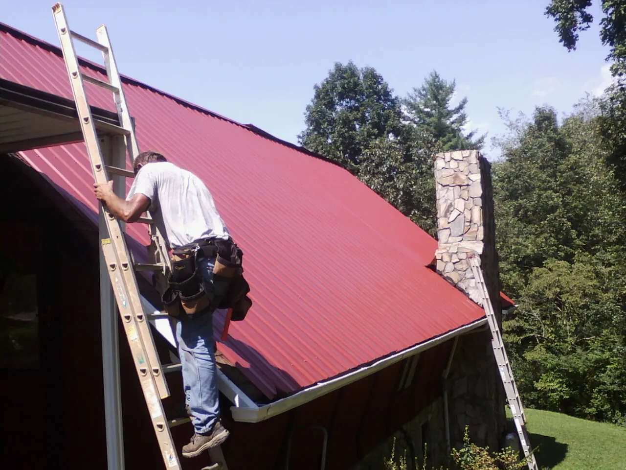 Roofer on a ladder beside a red metal roof and stone chimney — Soffit & Fascia Repair by Roofing Brothers in Bluemont